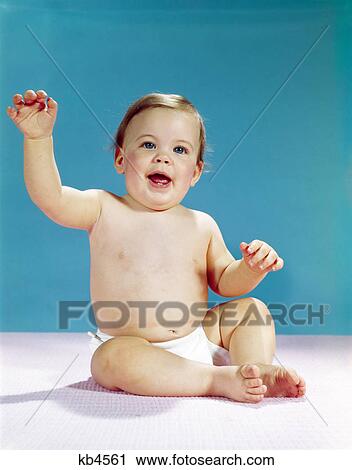Stock Image - 1960s happy smiling baby wearing diaper sitting and waving raised arm. Fotosearch
