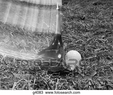 Stock Image - 1960s moving driver golf club hitting ball on tee in grass. Fotosearch