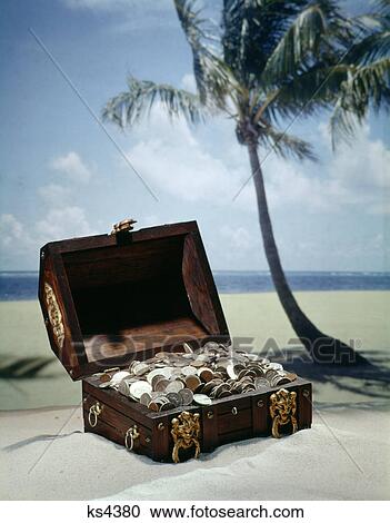 Stock Image - 1960s pirate treasure chest full coins on tropical beach. Fotosearch