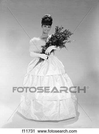 Stock Image - 1960s pretty young woman in evening dress at beauty pageant wearing fur stole and tiara holding bouquet of roses. Fotosearch
