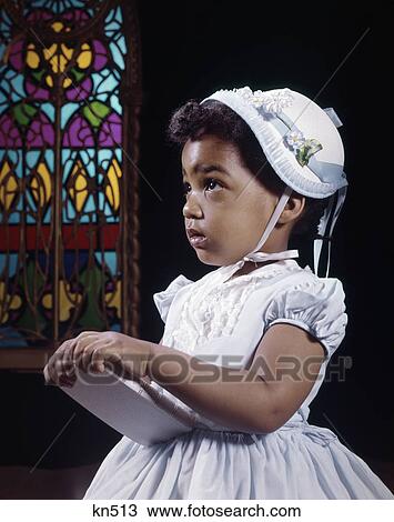 Stock Image - 1960s religious little african american girl wearing white hat and dress holding bible standing by stained glass window in church. Fotosearch