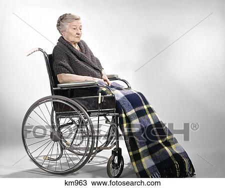Stock Image - 1960s sad senior woman sitting in wheelchair shawl on shoulders and a plaid lap blanket. Fotosearch