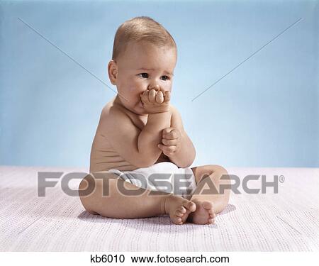 Stock Image - 1960s serious baby wearing cloth diaper putting fingers in mouth. Fotosearch