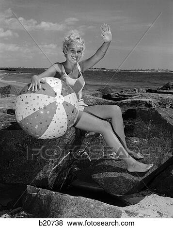 1960s smiling blond teenage girl sitting on rocky shore holding a beach ball waving View Large Photo Image Stock Photo - 1960s smiling blond teenage girl sitting on rocky shore holding a beach ball waving. Fotosearch