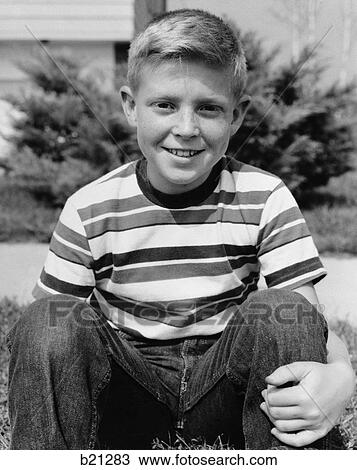 Stock Image - 1960s smiling boy sitting wearing blue jeans and striped jersey shirt looking at camera. Fotosearch