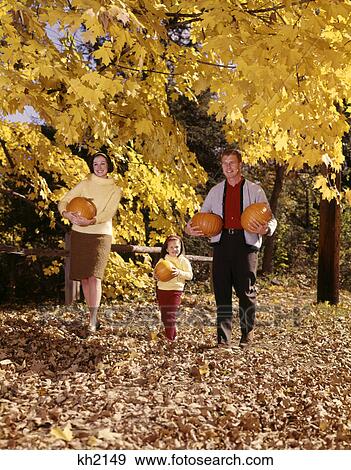 Stock Photo - 1960s smiling family trio father mother daughter walking in autumn woods carrying pumpkins. Fotosearch