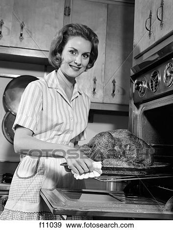 Stock Photo - 1960s smiling housewife removing thanksgiving roast turkey from oven. Fotosearch