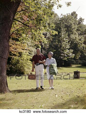 1960s smiling senior couple walking arm in arm carrying picnic basket View Large Photo Image Stock Image - 1960s smiling senior couple walking arm in arm carrying picnic basket. Fotosearch