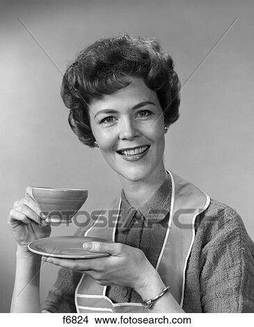 1960s smiling woman housewife wearing apron holding cup and saucer of coffee or tea looking at camera View Large Photo Image Picture - 1960s smiling woman housewife wearing apron holding cup and saucer of coffee or tea looking at camera. Fotosearch