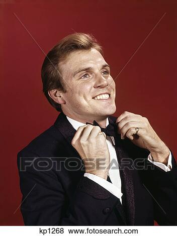 Stock Photo - 1960s smiling young man adjusting the bow tie of his tuxedo. Fotosearch