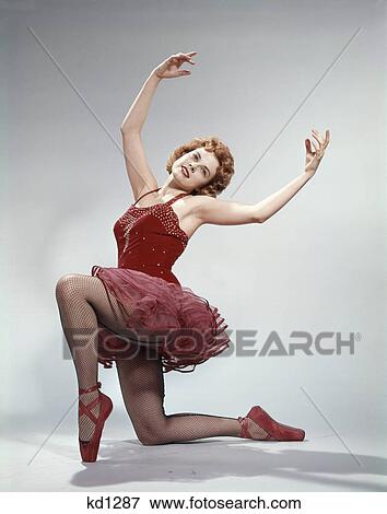 Stock Photo - 1960s teen girl woman red velvet ballet costume pink net tutu kneeling arms above attitude position. Fotosearch