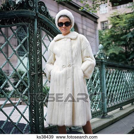 Stock Photo - 1960s woman wearing white mink fur coat hat sunglasses by wrought iron gate. Fotosearch