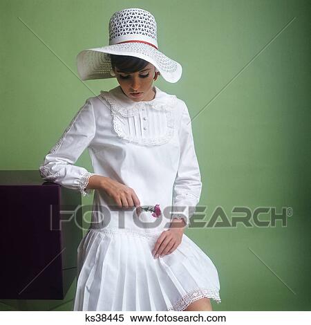 Stock Photography - 1960s young woman white floppy straw hat white dress ruffles pleated skirt head lowered holding pink flower bride. Fotosearch