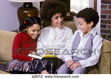 Stock Image - 1970s 1980s african american mother sitting on couch with son and daughter reading together. Fotosearch
