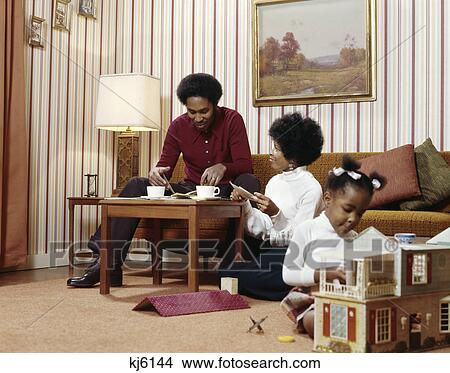 1970s african american family mother father at coffee table with daughter playing doll house in living room View Large Photo Image Picture - 1970s african american family mother father at coffee table with daughter playing doll house in living room. Fotosearch