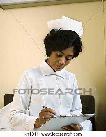 Stock Image - 1970s african american woman nurse filling out medical chart. Fotosearch