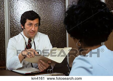Stock Image - 1970s african american woman patient consulting with caucasian man doctor in office. Fotosearch