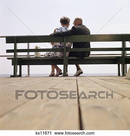 1970s back view of senior couple sitting on boardwalk bench overlooking ocean beach View Large Photo Image Stock Image - 1970s back view of senior couple sitting on boardwalk bench overlooking ocean beach. Fotosearch