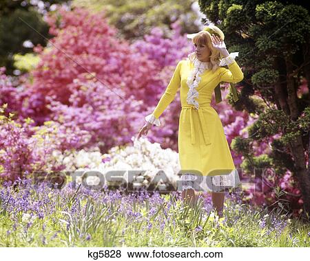 Stock Photo - 1970s blonde woman yellow dress straw hat spring landscape pink white azaleas blossoms fashion outdoor. Fotosearch