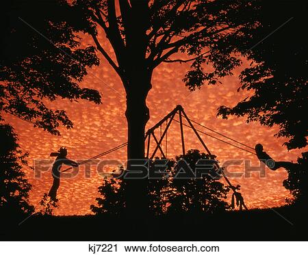 Stock Image - 1970s composite image silhouetted children playing on swing set by large tree at sunset. Fotosearch