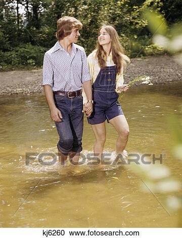 1970s couple man woman water walking lake pond holding hands romance love jeans overalls happy smile View Large Photo Image Stock Image - 1970s couple man woman water walking lake pond holding hands romance love jeans overalls happy smile. Fotosearch