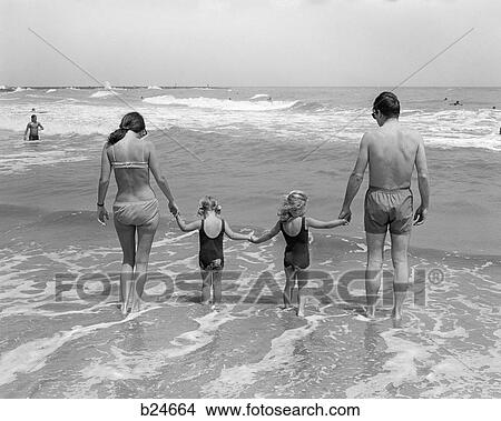 Picture - 1970s family on vacation at ocean beach holding hands walking on sand in surf. Fotosearch