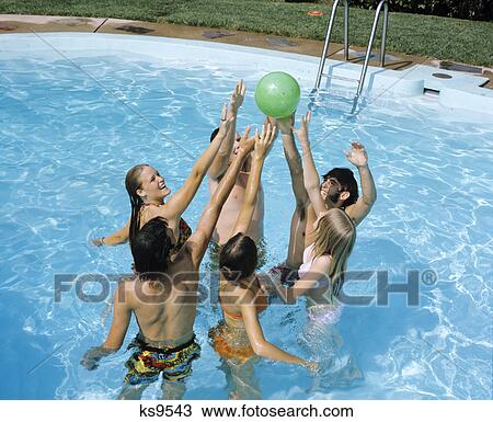 Stock Image - 1970s group teens boys girls in pool jumping after ball pool party. Fotosearch