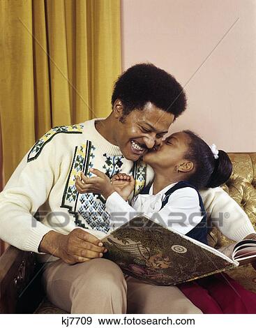 1970s happy african american little girl kissing her smiling father as they read a book together View Large Photo Image Stock Photo - 1970s happy african american little girl kissing her smiling father as they read a book together. Fotosearch