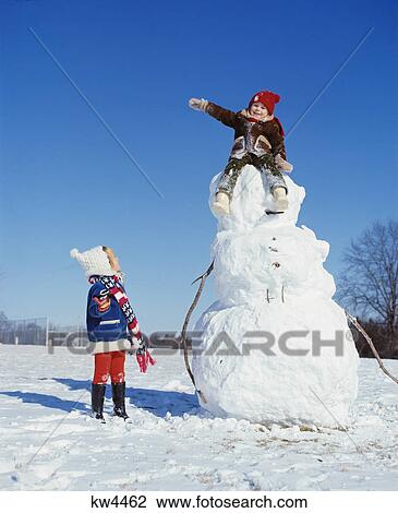 Stock Image - 1970s kids playing in snow girl looking up at boy waving sitting top snowman. Fotosearch