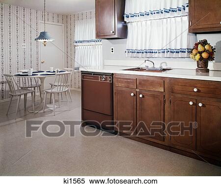 Stock Photography - 1970s kitchen and dining area with dark wood cabinets and blue and white curtains. Fotosearch