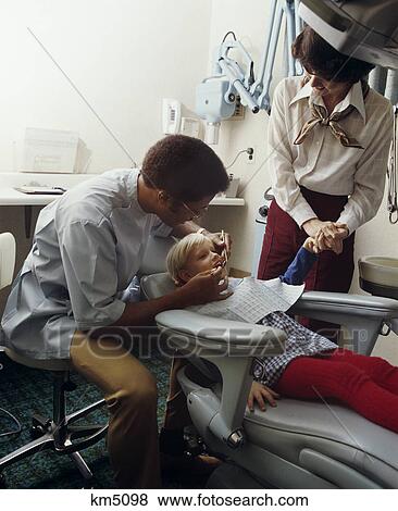 Stock Photo - 1970s man african american dentist working on little blond girl patient with woman mother holding her hand. Fotosearch