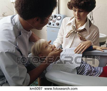 Stock Photo - 1970s man african american dentist with little blond girl patient with woman mother standing by. Fotosearch