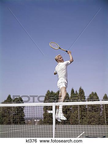 1970s man playing tennis jumping in mid air swinging racket to hit ball action recreation exercise View Large Photo Image Stock Photograph - 1970s man playing tennis jumping in mid air swinging racket to hit ball action recreation exercise. Fotosearch