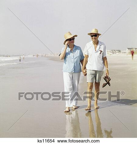 Picture - 1970s senior couple walking beach holding hands wearing straw hats. Fotosearch