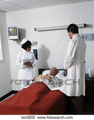 Stock Image - 1970s senior man in hospital bed under red blanket talking with caucasian doctor and african american nurse at his bedside. Fotosearch
