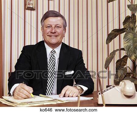 1970s smiling businessman sitting at desk writing papers folders View Large Photo Image Stock Photograph - 1970s smiling businessman sitting at desk writing papers folders. Fotosearch