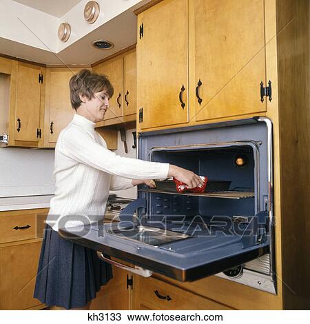 1970s smiling housewife in kitchen taking baking dish out of wall mounted oven View Large Photo Image Stock Image - 1970s smiling housewife in kitchen taking baking dish out of wall mounted oven. Fotosearch