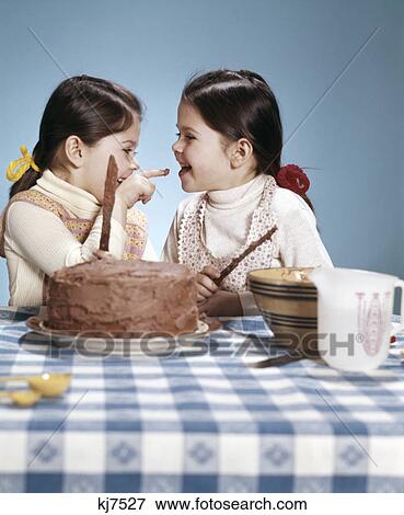 Stock Photo - 1970s two sisters twins smiling frosting chocolate cake tasting frosting baking cooking. Fotosearch