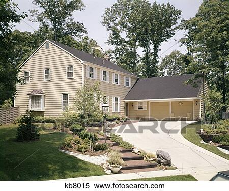 Stock Photography - 1970s two story yellow house white shutters with two car garage curved driveway landscaped lawn suburban. Fotosearch