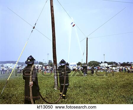 Stock Image - 1970s volunteer fireman's picnic two teams fire fighters battle push barrel water hoses contest. Fotosearch