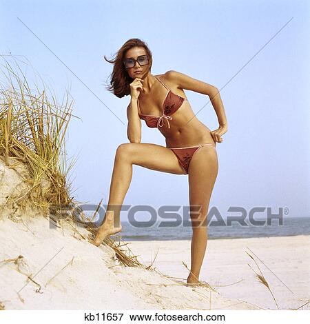 1970s woman wearing two piece string bikini bathing suit sunglasses posing by grassy sand dune at beach View Large Photo Image Stock Photo - 1970s woman wearing two piece string bikini bathing suit sunglasses posing by grassy sand dune at beach. Fotosearch