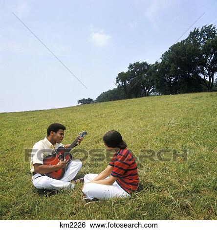 1970s young african american couple sitting on grassy hill man playing acoustic guitar View Large Photo Image Stock Photograph - 1970s young african american couple sitting on grassy hill man playing acoustic guitar. Fotosearch