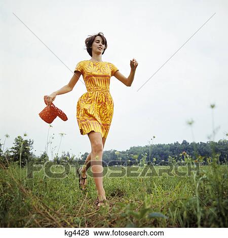 Stock Photo - 1970s young woman wearing colorful print dress running in field carrying red straw hat. Fotosearch