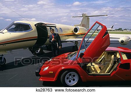 Stock Image - 1980s business man standing between lamborghini automobile and lear jet. Fotosearch