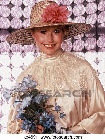 Stock Image - 1980s portrait of a woman wearing a straw hat with a pink flower holding an artificial bouquet. Fotosearch