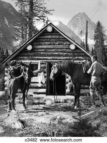 Stock Image - 1920S 1930S Couple In Front Of Log Cabin Standing By Horses With Saddles Both Wearing Jodhpurs Assiniboine Canada. Fotosearch