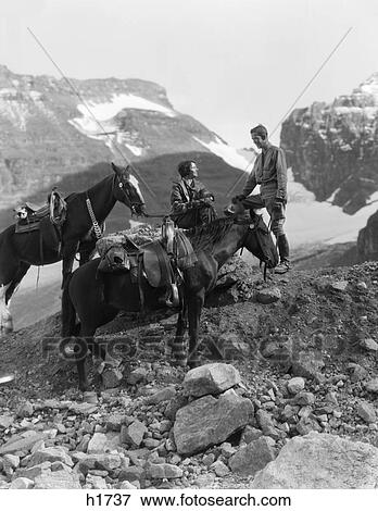Stock Photo - 1920S 1930S Couple Man Woman Wearing Riding Gear Jodhpurs Boots Spurs Sitting Standing On Large Rock By Two Horses. Fotosearch