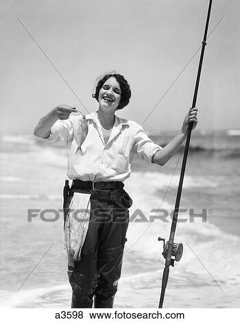 Stock Photo - 1920S 1930S Smiling Woman Standing In Ocean Surf Wearing Rubber Waders Holding Fish And Fly Fishing Rod. Fotosearch