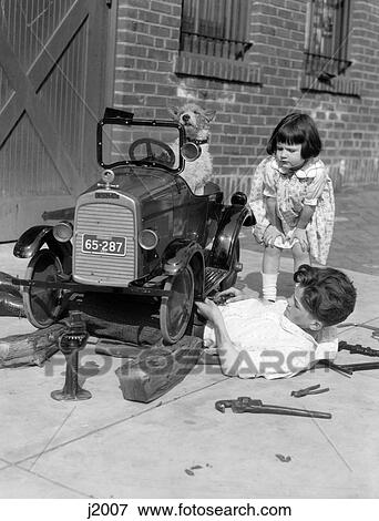 Stock Photo - 1920S Little Girl Leaning Down Watching Boy Working Under Toy Car With Dog Behind Wheel. Fotosearch