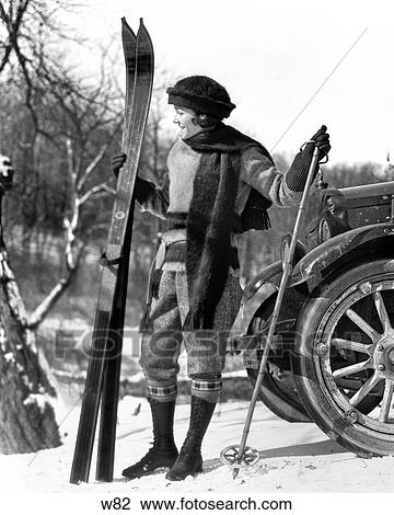Stock Image - 1920S Woman In Scarf Hat And Boots Standing In Front Of Car Holding Wood Skis And Bamboo Ski Poles. Fotosearch